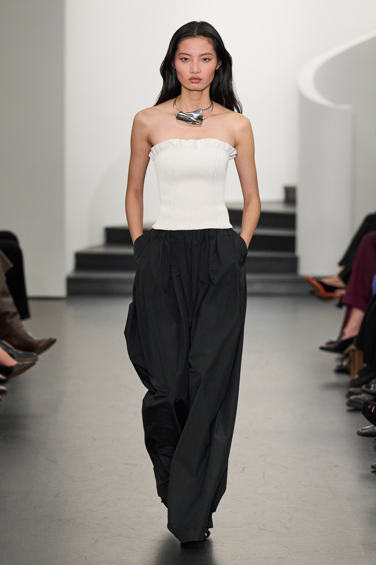 Female model on a runway wearing a white strapless ribbed top and oversized black trousers, accessorized with a chunky silver necklace.