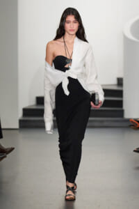Runway model in a black strapless dress with a white shirt tied at the waist, walking in a fashion show with a layered necklace and clutch.