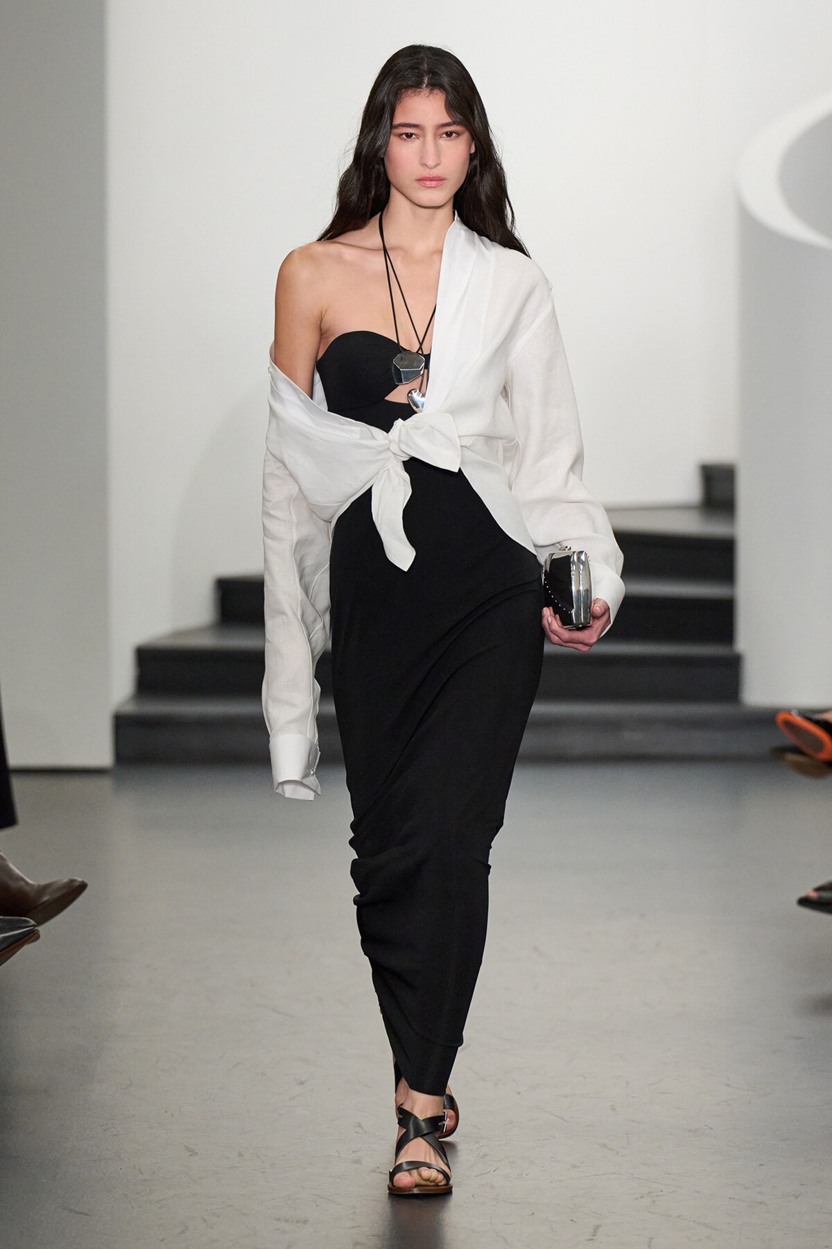 Runway model in a black strapless dress with a white shirt tied at the waist, walking in a fashion show with a layered necklace and clutch.