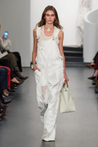 Model on a runway wearing white patchwork overalls over a mesh tank top, carrying a white textured handbag.
