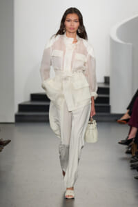 Model walks a white-on-white runway look: sheer blouse with large pockets, paired with a belted, oversized utility-style trouser set and a small white handbag.