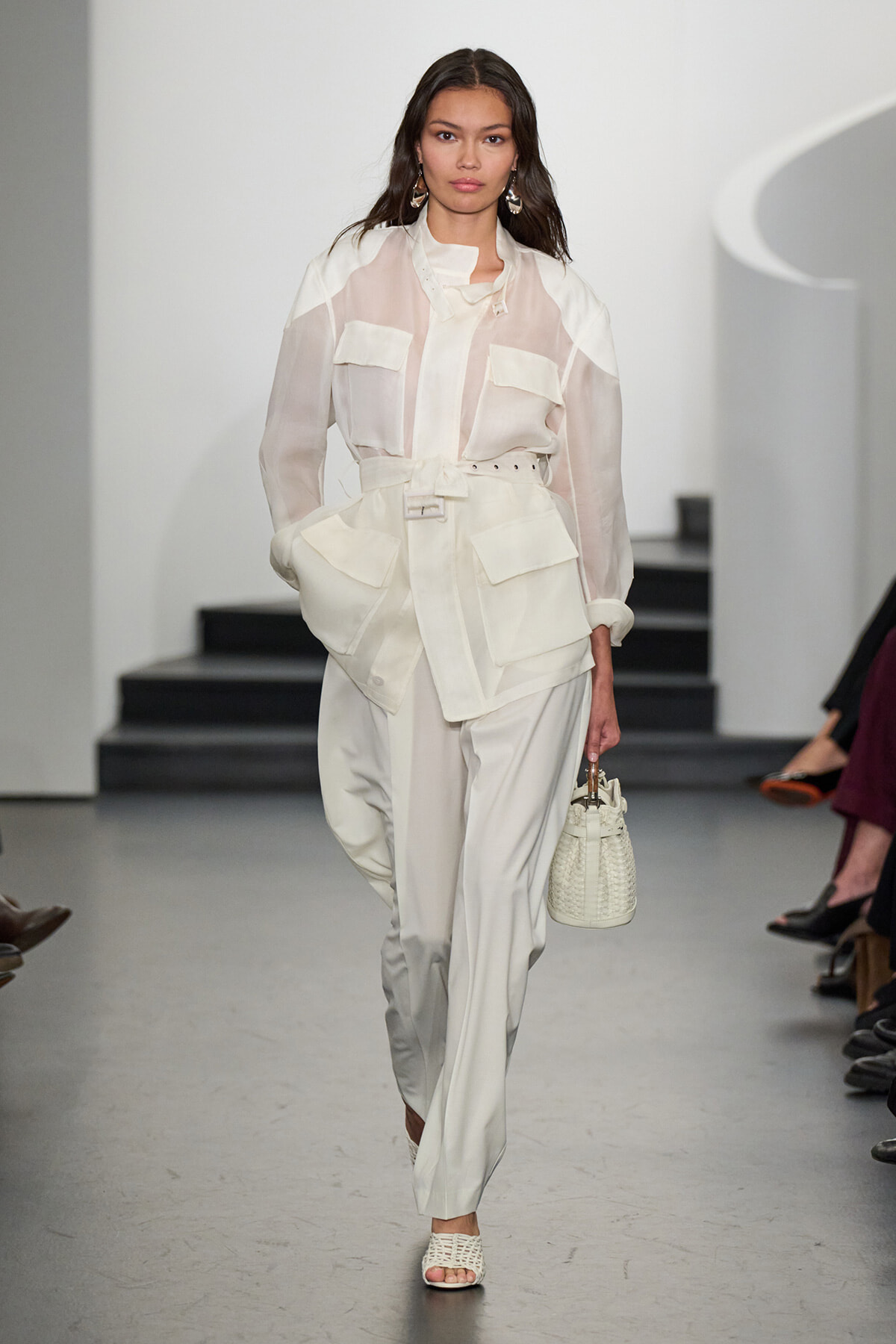 Model walks a white-on-white runway look: sheer blouse with large pockets, paired with a belted, oversized utility-style trouser set and a small white handbag.