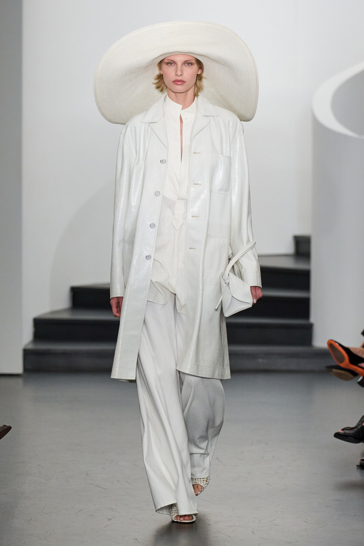 Model walks a runway in an all-white ensemble: oversized wide-brim hat, long coat, wide-leg pants, and a white handbag.