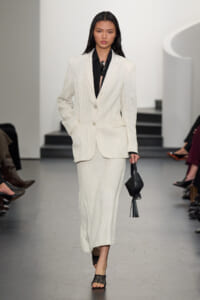 Model walking the runway in a white pinstripe suit (blazer and wide-leg pants) with a black blouse and black tassel handbag.
