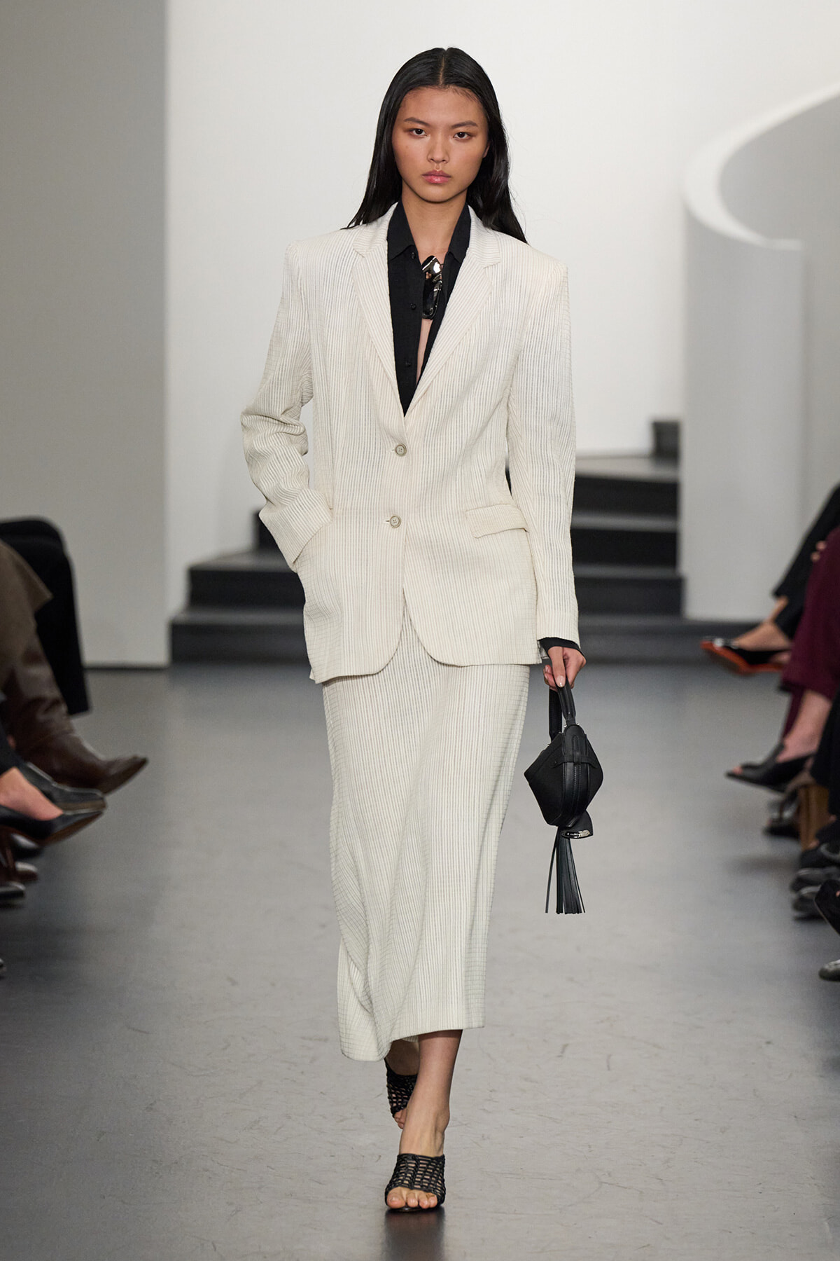 Model walking the runway in a white pinstripe suit (blazer and wide-leg pants) with a black blouse and black tassel handbag.
