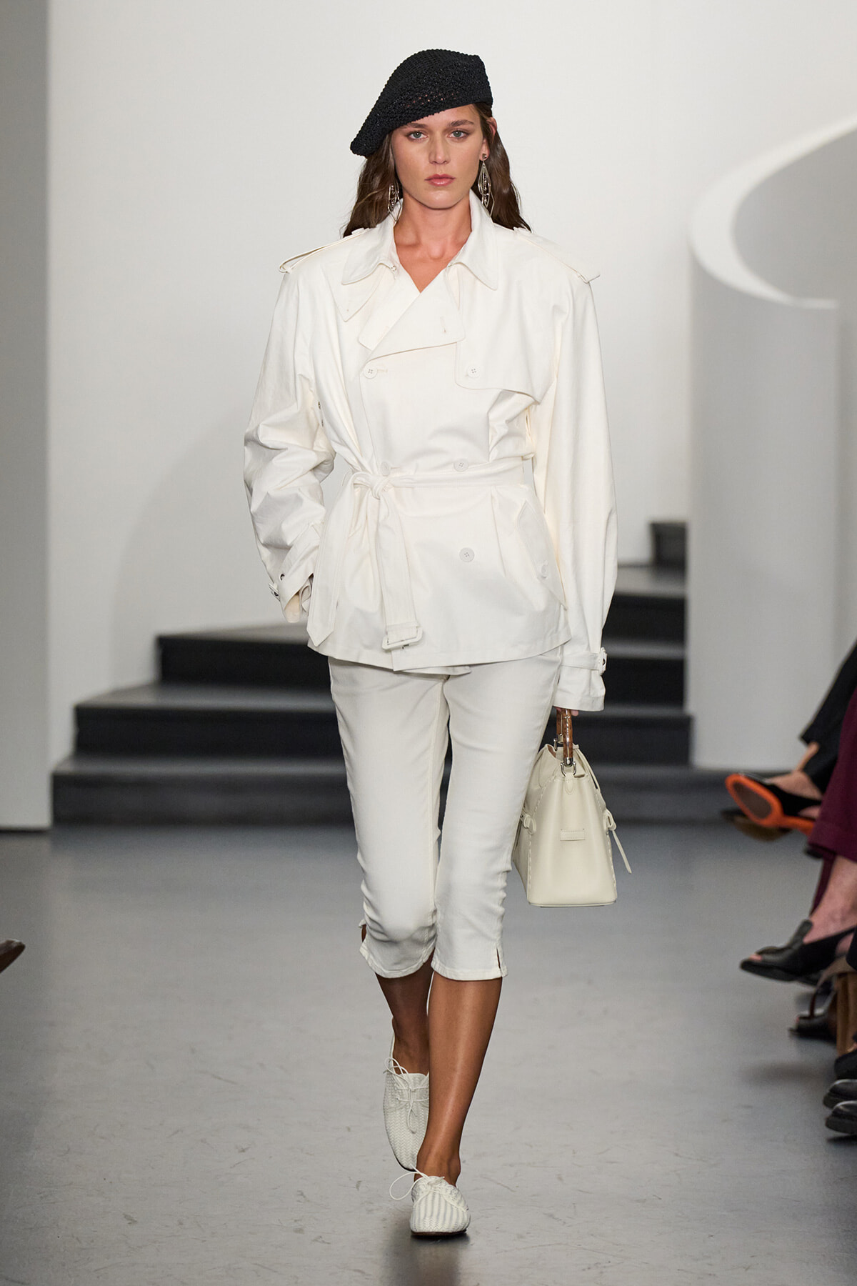 Model walking a runway in an all-white outfit: belted trench with rolled sleeves, cropped white pants, white loafers, a black knit beret, and a white handbag.