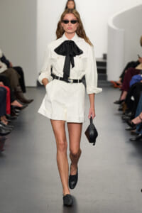 Model walks the runway in a white utility romper with rolled sleeves, a large black neck bow, black belt, and small black bag, wearing dark sunglasses.