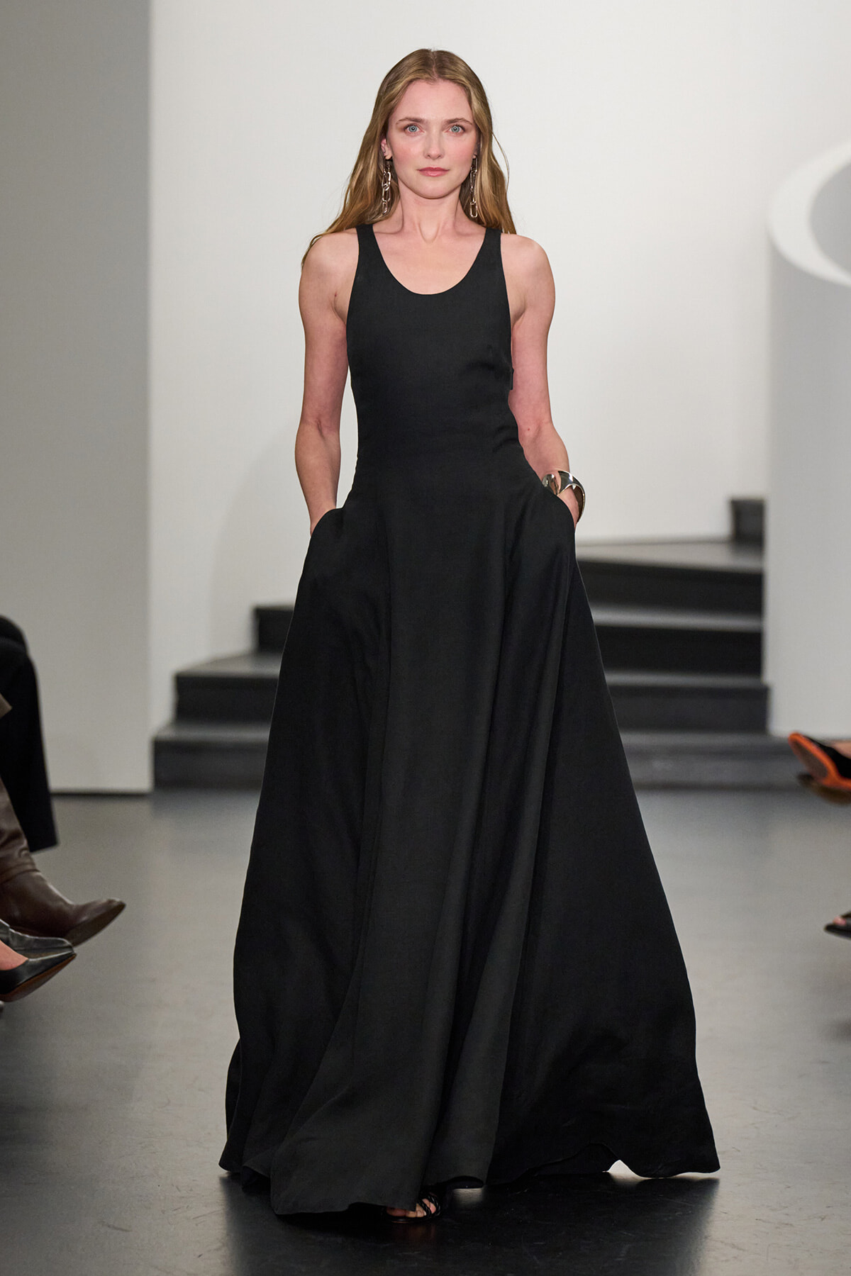 Model walking a fashion runway in a floor-length black sleeveless dress with pockets."
