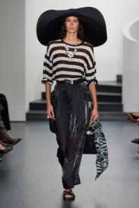 Model on runway wearing a large black floppy hat, beige‑black striped sweater, glossy black pants, statement necklace, and a black bag with a patterned scarf.