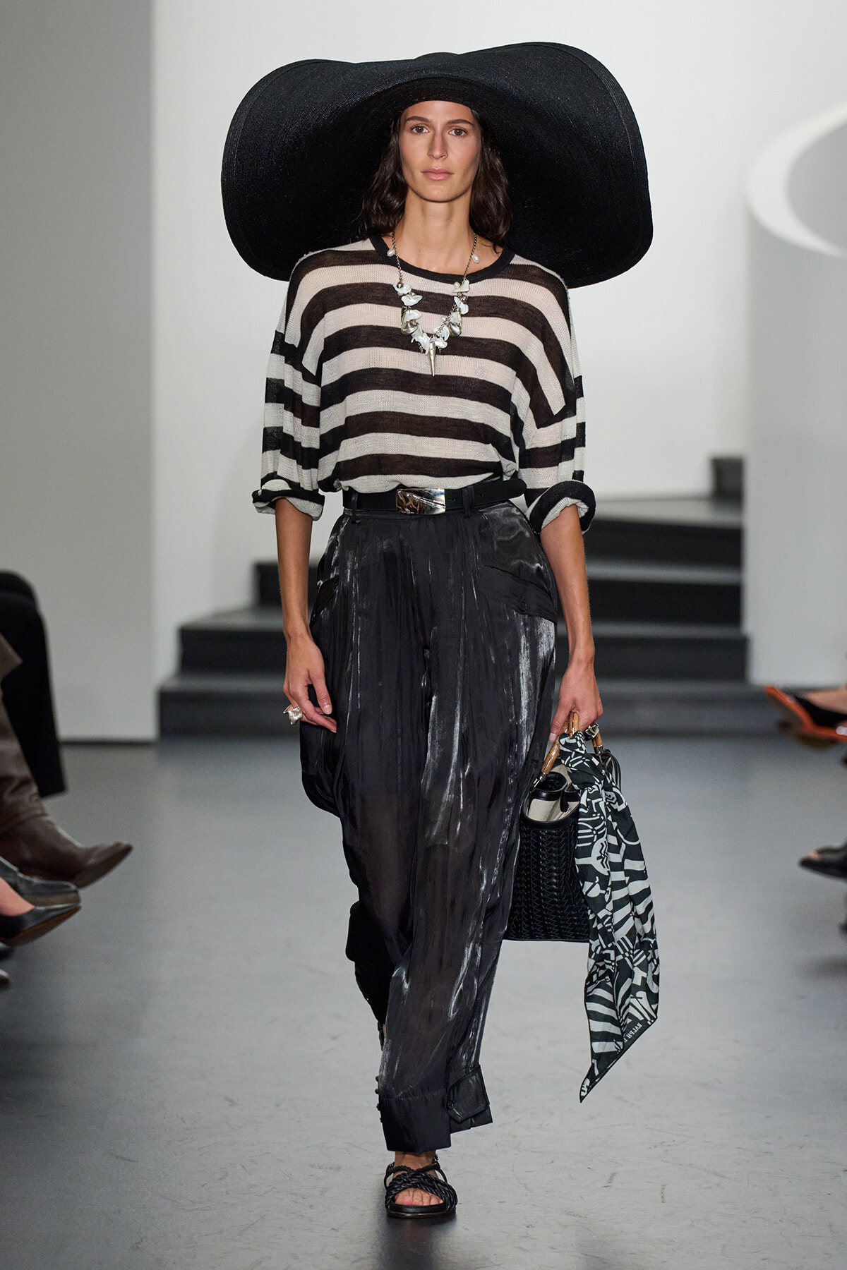 Model on runway wearing a large black floppy hat, beige‑black striped sweater, glossy black pants, statement necklace, and a black bag with a patterned scarf.