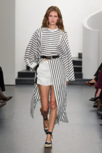 Model walking a runway in a black-and-white striped outfit: long striped cape over a buttoned top and white shorts, with a black belt and espadrille sandals.