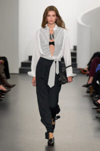 Model walking the runway in a white striped blouse tied at the waist over a black bralette, paired with black wide-leg pants and a black handbag.