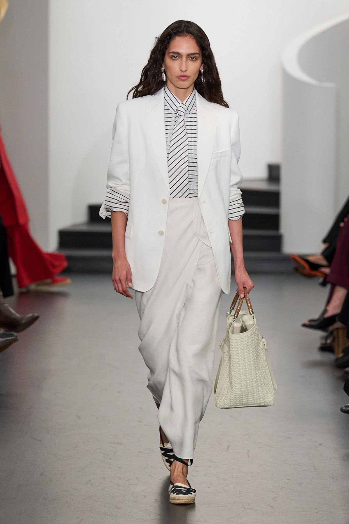 Female model walking a fashion runway in a tailored white suit with a striped blouse, carrying a woven white handbag.