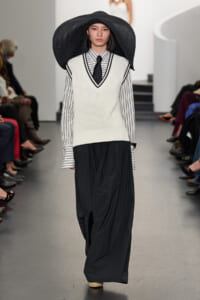 Model walking the runway in a wide-brim black hat, white V-neck knit vest over a striped shirt with a black tie, and flowing black pants.