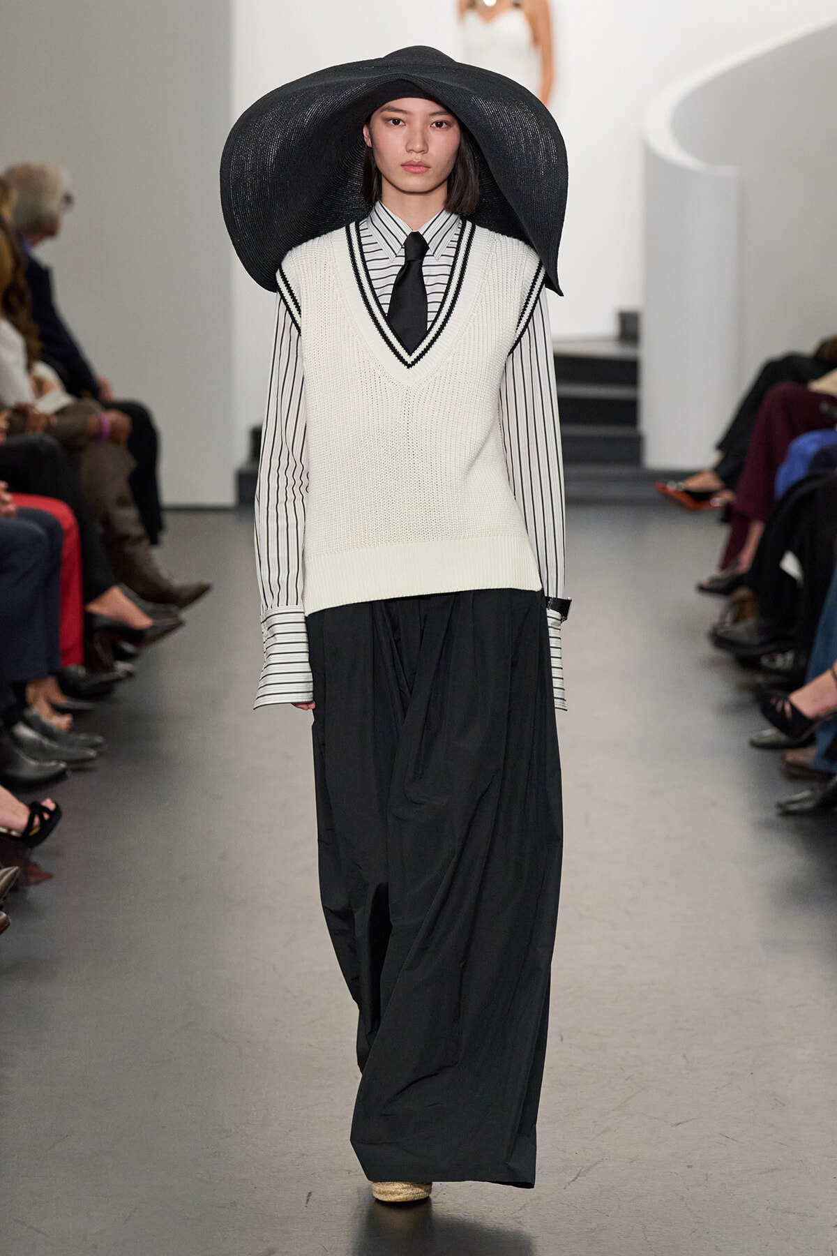 Model walking the runway in a wide-brim black hat, white V-neck knit vest over a striped shirt with a black tie, and flowing black pants.