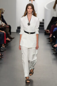 Model walks a fashion runway in an all-white tailored blazer and cargo-style pants with a black belt and black strappy sandals, wearing a large pendant necklace and earrings.