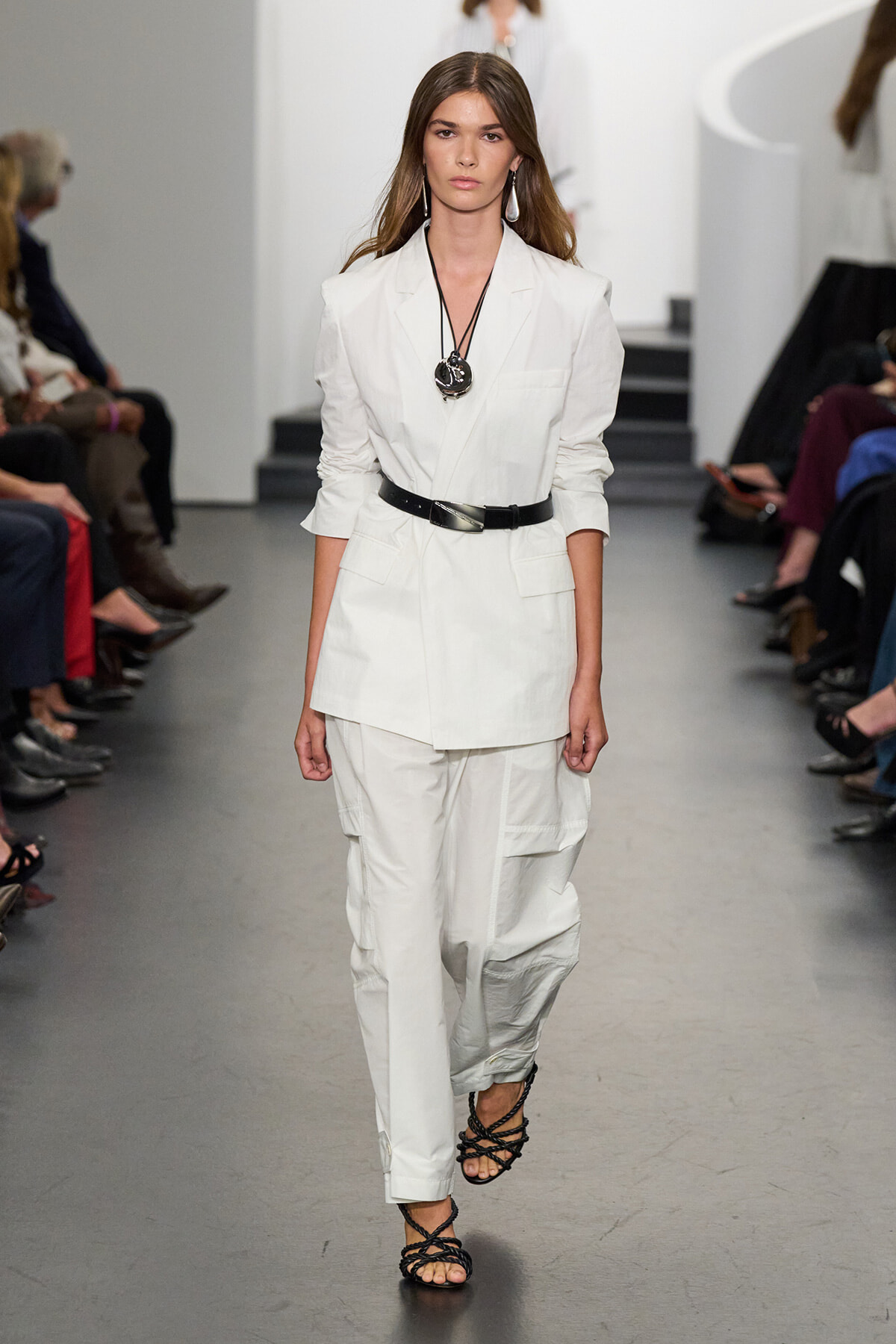 Model walks a fashion runway in an all-white tailored blazer and cargo-style pants with a black belt and black strappy sandals, wearing a large pendant necklace and earrings.