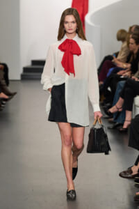 Model walking a runway in a white sheer blouse with a red necktie bow, black mini skirt, and black woven handbag.
