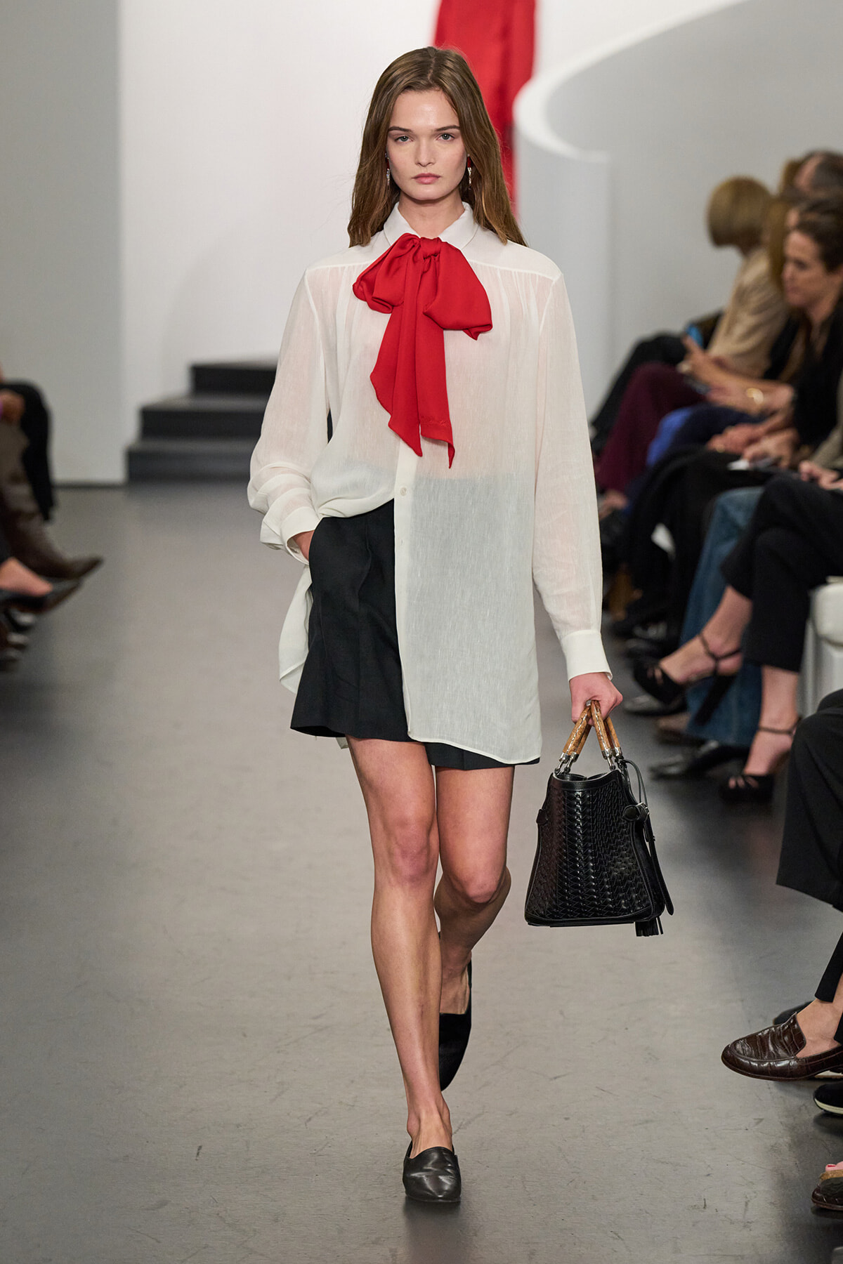 Model walking a runway in a white sheer blouse with a red necktie bow, black mini skirt, and black woven handbag.