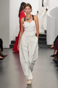 Model walking the runway in a white pinstripe sleeveless vest and wide-leg pants with a black halter strap, silver necklace, and white sandals.
