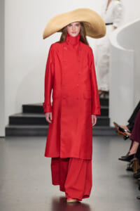 Model walks a fashion-show runway wearing a bright red double-breasted trench coat and an oversized straw hat.