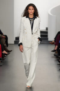Runway model wearing a white pinstripe double-breasted suit with black lapels and a striped top, hands in pockets.
