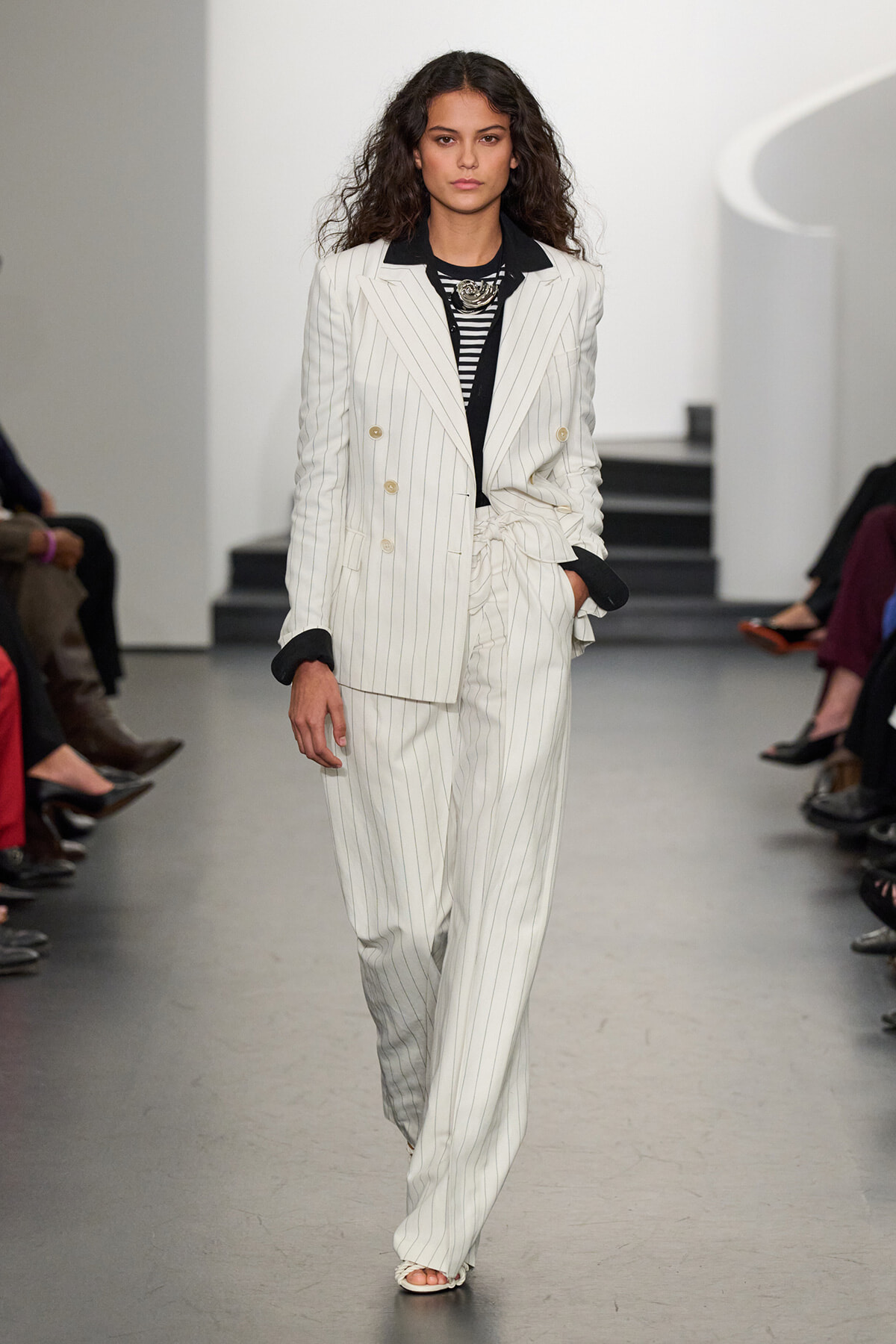 Runway model wearing a white pinstripe double-breasted suit with black lapels and a striped top, hands in pockets.
