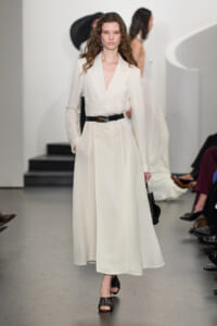 Runway model in a cream double-breasted coat with a black belt, holding a small black bag as she walks the catwalk.