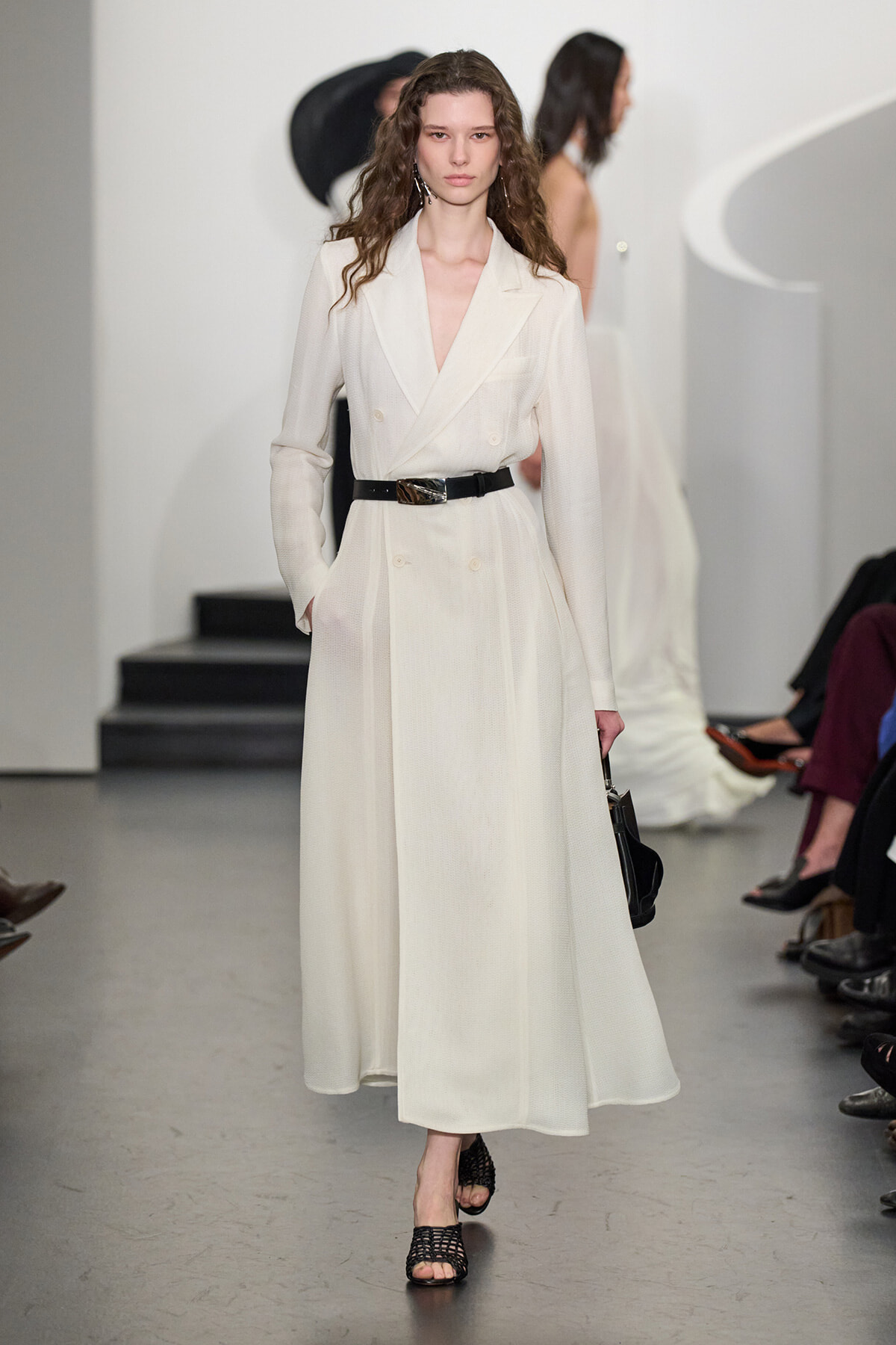 Runway model in a cream double-breasted coat with a black belt, holding a small black bag as she walks the catwalk.