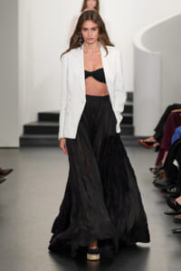 Model on a runway wearing a white blazer over a black bandeau top and a long, crinkled black skirt.