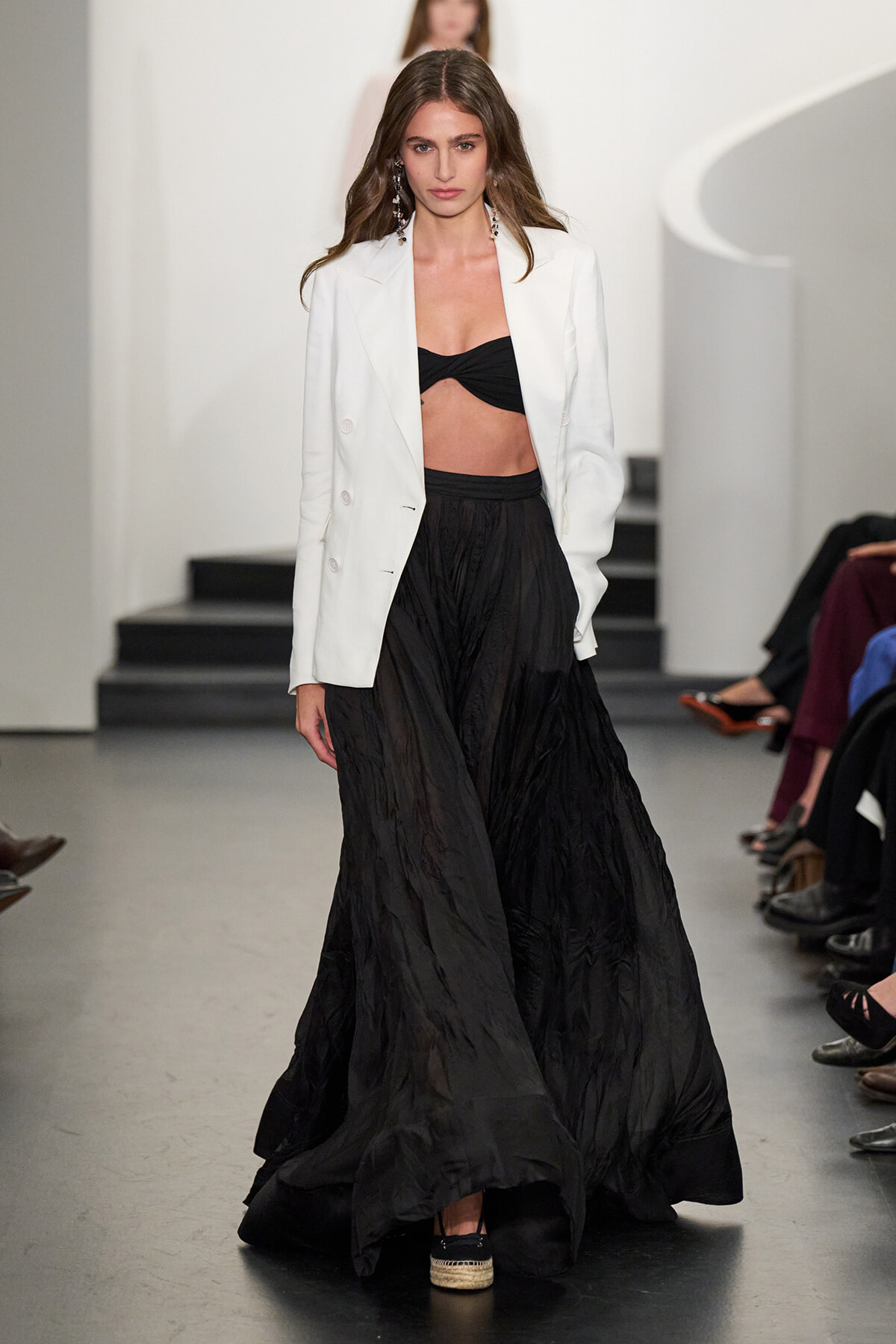 Model on a runway wearing a white blazer over a black bandeau top and a long, crinkled black skirt.