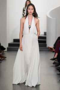 Female model walks the runway in a white halter gown with a plunging neckline and a long flowing skirt, accessorized by a large silver spoon-shaped pendant on a black cord.