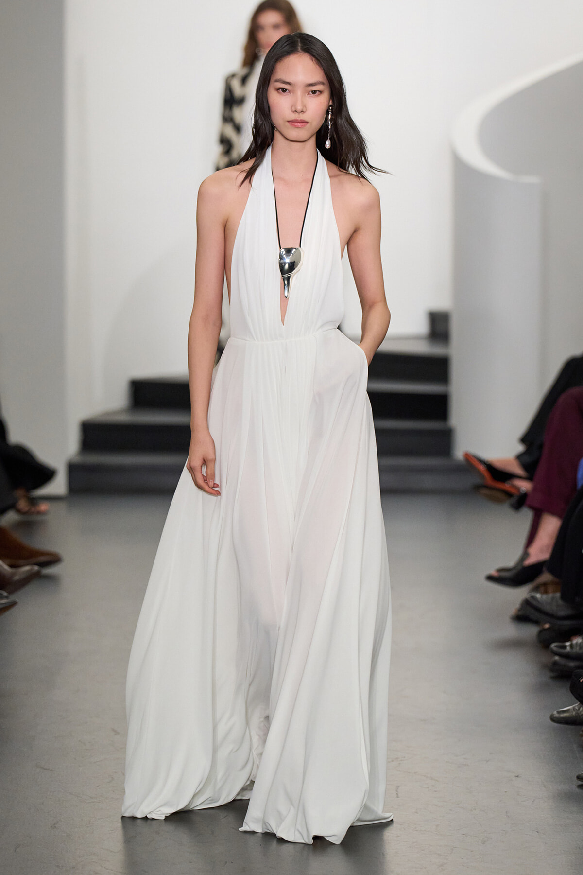 Female model walks the runway in a white halter gown with a plunging neckline and a long flowing skirt, accessorized by a large silver spoon-shaped pendant on a black cord.