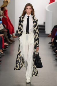 Model walking a runway in a black-and-cream geometric coat over a white shirt and wide white trousers, carrying a black handbag.