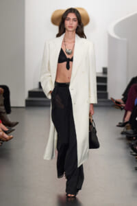 Model on a runway wearing a long white blazer over a black bralette and black wide-leg pants, with a chunky silver necklace and a black handbag.