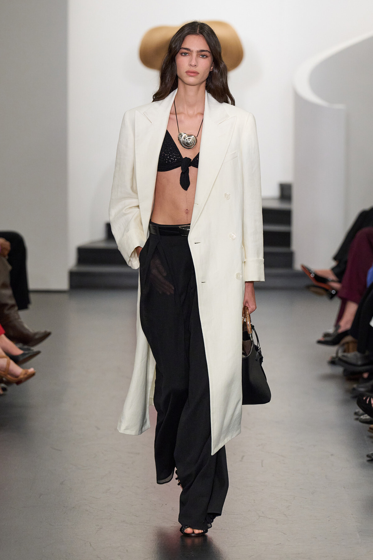 Model on a runway wearing a long white blazer over a black bralette and black wide-leg pants, with a chunky silver necklace and a black handbag.