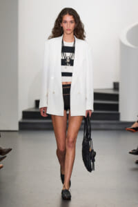 Model on a runway wearing a white double-breasted blazer over a black-and-white striped crop top, black crochet shorts, and a black woven bag.