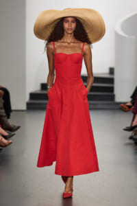 Model walking a runway in a bright red, structured midi dress with pockets and a wide straw hat.