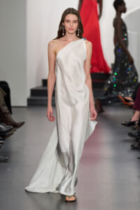Model walking a runway in a white one-shoulder satin gown with drop earrings and neutral makeup; minimalist elegance.