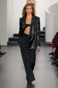 Model walking runway in a black leather blazer, black bralette, and wide-leg trousers with layered necklaces and a ring.