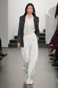 Model walking a runway in a black sequined blazer, white shirt, and wide-leg white trousers with a waist tie.