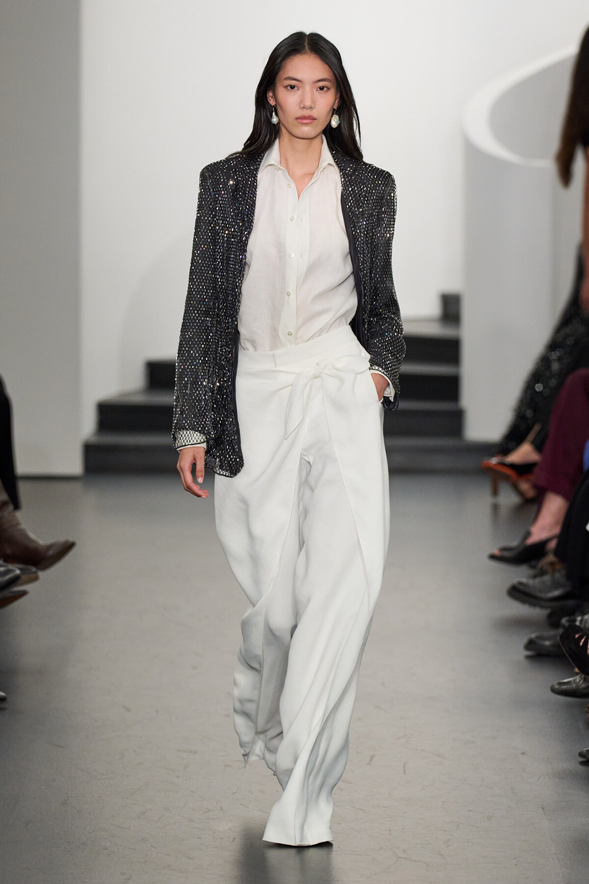 Model walking a runway in a black sequined blazer, white shirt, and wide-leg white trousers with a waist tie.
