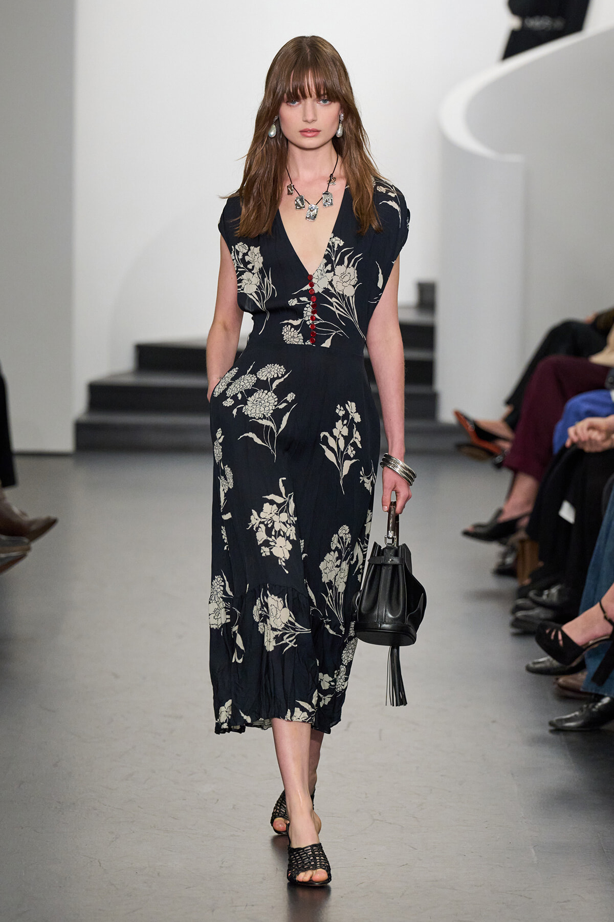 Model walking runway in a navy floral midi dress with red button accents, holding a black handbag and wearing statement jewelry.
