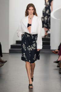 Runway model wearing a white oversized shirt open over a black bra and a navy floral midi skirt with a black belt and sandals.