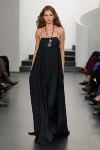 Model walking runway in a loose black spaghetti-strap gown, layered, floor-length with pockets visible and a geometric pendant necklace