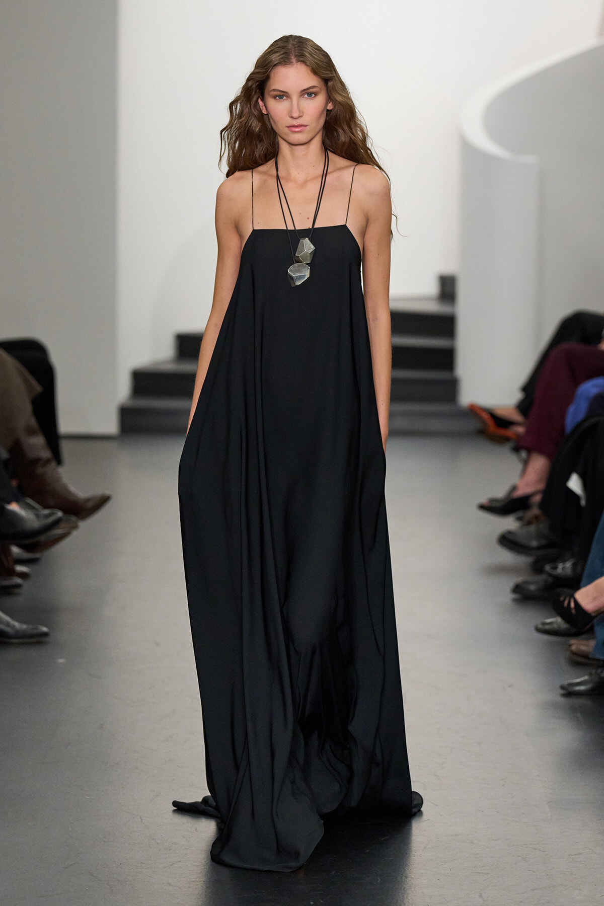 Model walking runway in a loose black spaghetti-strap gown, layered, floor-length with pockets visible and a geometric pendant necklace