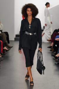 Model walking a fashion show wearing a black button-down blouse, black cropped pants, a patterned belt, and layered necklace with a black woven bag.",