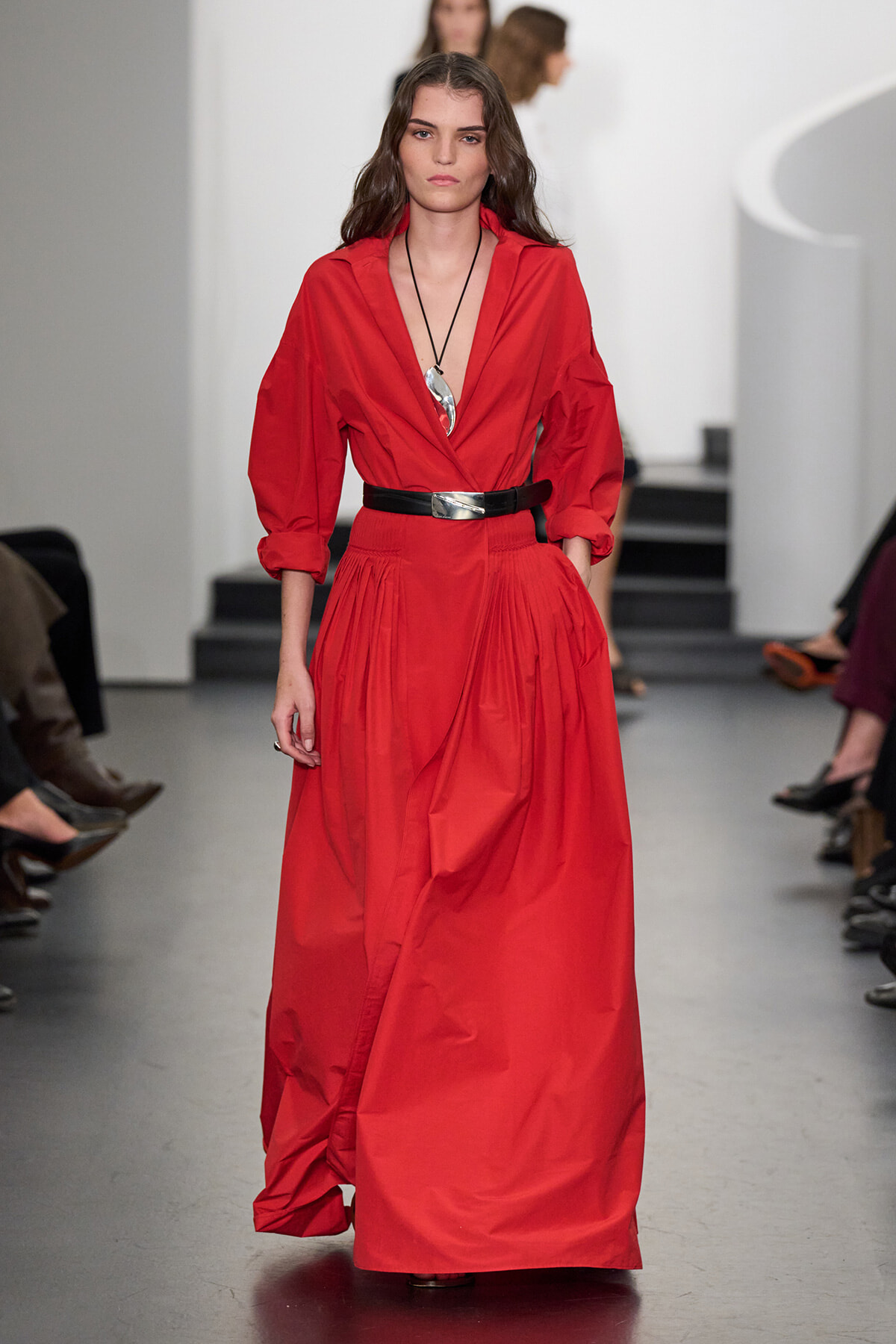 Model walking the runway in a long red dress with a black belt and a V-neck, wearing a silver pendant necklace.