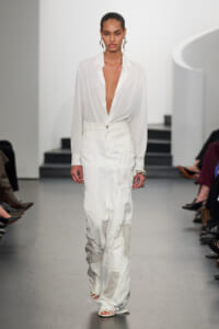 Model walking a fashion runway in a white sheer button-down blouse with a deep V neckline and high-waisted patchwork white trousers.