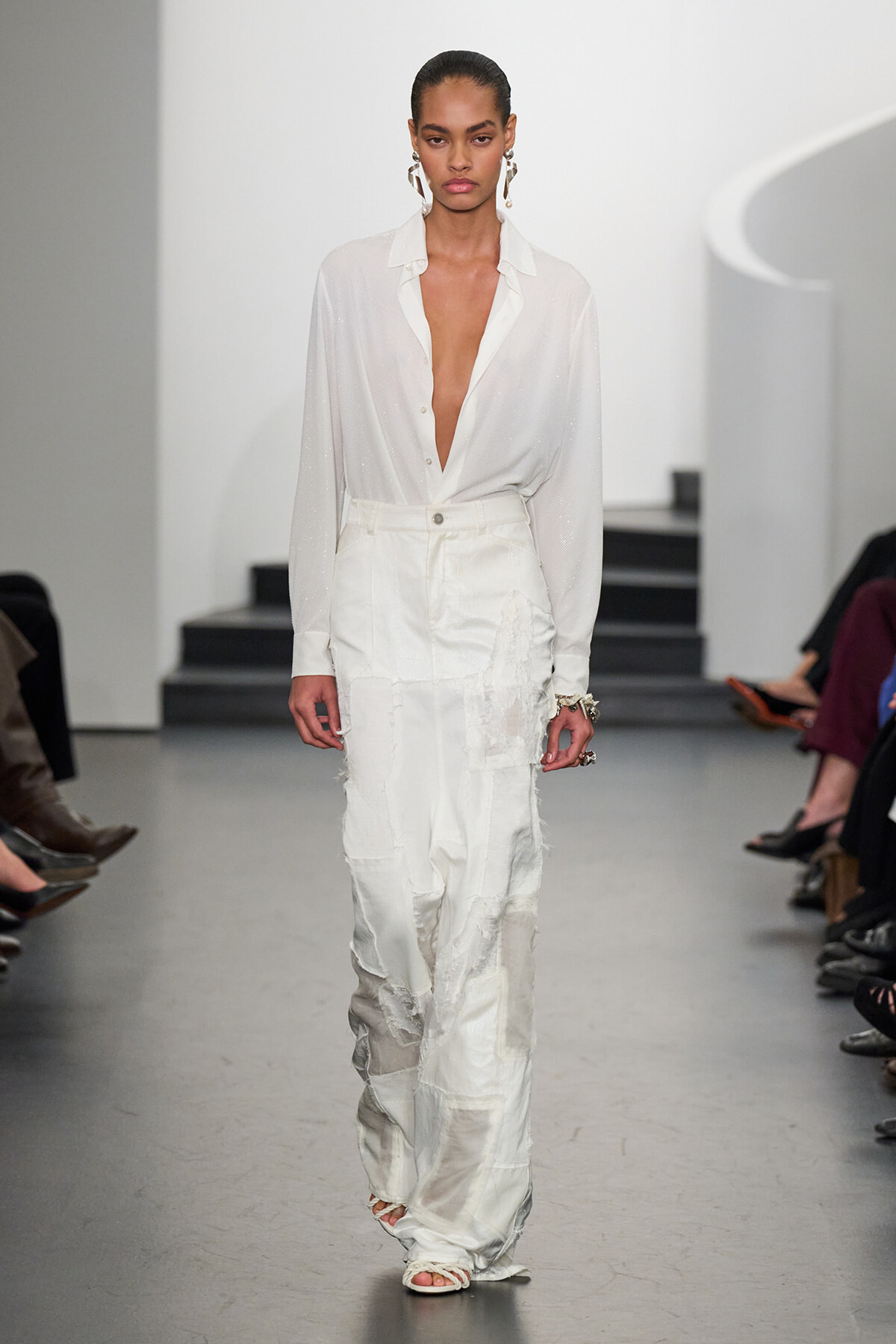 Model walking a fashion runway in a white sheer button-down blouse with a deep V neckline and high-waisted patchwork white trousers.